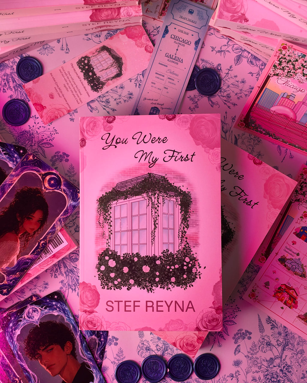 A pink book or booklet titled "You Were My First" by Stef Reyna is centered on a pink floral surface, surrounded by matching tickets, cards, seals, and photo inserts in a coordinated flat-lay arrangement.